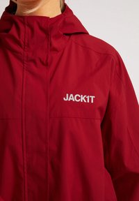 Personne portant une veste imperméable rouge avec une capuche et le logo réfléchissant "JACK1T" sur la poitrine, sur un fond clair.