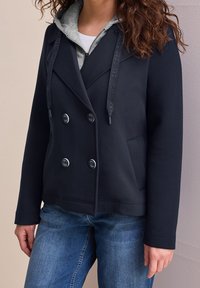 Femme portant une veste croisée bleu marine sur un sweat à capuche gris avec des cordons à logo, et un jean bleu, debout contre un mur de couleur claire.