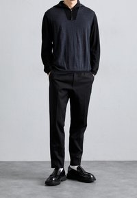 Pull noir avec un col à fermeture éclair, présentant un panneau gris sur la poitrine. Associé à un pantalon noir et des chaussures noires polies.