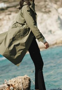 Olive zielony płaszcz trencz z zaznaczoną talią, dużymi guzikami i kapturem, zestawiony z czarnymi dopasowanymi spodniami, przedstawiony na tle nadmorskim.