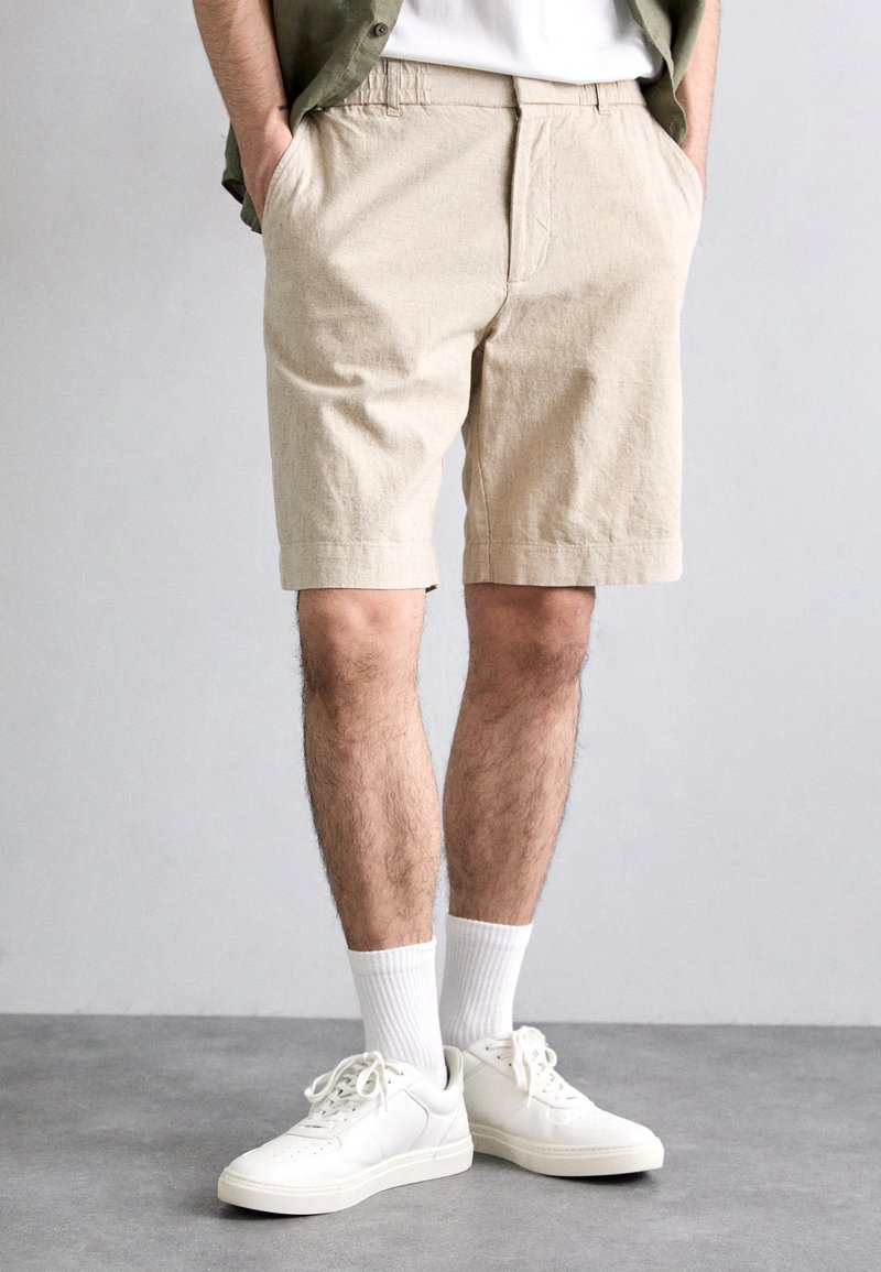 Shorts en lin beige avec une coupe décontractée, des poches latérales et une texture lisse. Portés avec des baskets blanches et des chaussettes blanches côtelées.