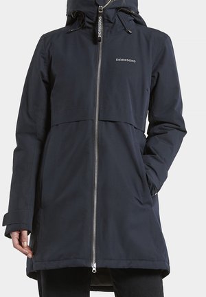 Abrigo impermeable Didriksons de color azul marino con capucha, cremallera completa en el frente y logo en el pecho, usado por una persona con las manos parcialmente visibles.