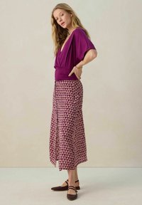 Haut violet à manches courtes enroulé associé à une longue jupe à motifs avec un design floral. Le mannequin porte des chaussures plates marron foncé.