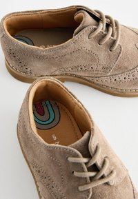 Chaussures en daim beige avec un bout arrondi, un laçage, des détails en brogue et une semelle en caoutchouc marron. L'intérieur présente une semelle intérieure à motif multicolore.