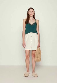 Top sem mangas em teal com rendas, combinado com uma saia branca de botão. Acessórios incluídos: uma bolsa tecida e sapatos slip-on bege.