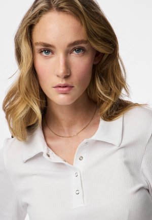 Tânără cu păr blond ondulat, ochi albaștri, purtând o cămașă albă cu dungi, cu nasturi capsa și un lanț subțire de aur, expresie neutră.