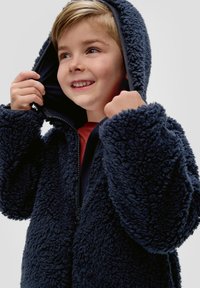 Marineblauwe nepbontjas met capuchon, met een ritssluiting aan de voorkant en elastische manchetten. De textuur is donzig en het is over een rode top gedragen.