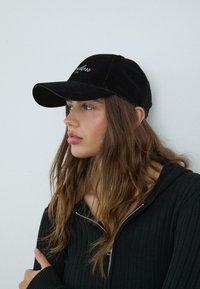 Black velvet baseball cap with a curved brim, featuring embroidered text on the front. The individual has long, wavy hair and wears a dark hoodie.