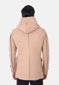 Cappotto di lana beige con cappuccio, caratterizzato da cuciture nere, due tasche laterali e un design aderente. La vista posteriore mostra la struttura e la texture del materiale.