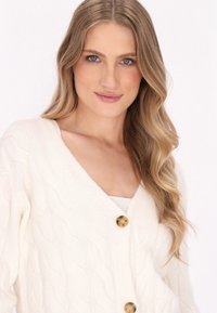 Witte kabelgebreide cardigan met V-hals, voorzien van grote gouden knopen. Zachte textuur met golvende patroon details door de stof.