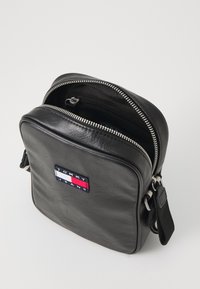 Čierna kožená crossbody taška so zipsom, obdĺžnikového tvaru, s výraznou nášivkou obsahujúcou modré, červené a biele logo.