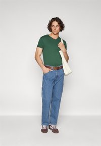 Green v-neck t-shirt, blue denim cargo pants, brown leather belt, and brown sneakers with white laces. Natural fabric tote bag slung over shoulder.