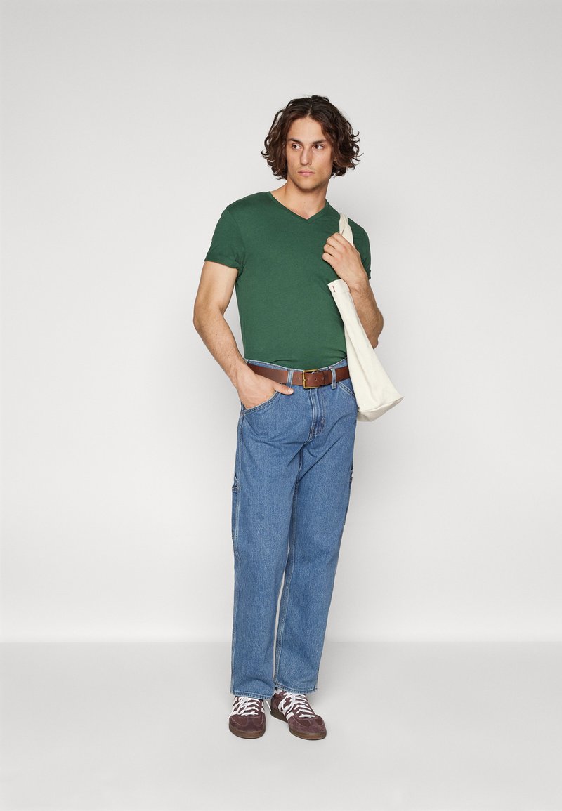 Green v-neck t-shirt, blue denim cargo pants, brown leather belt, and brown sneakers with white laces. Natural fabric tote bag slung over shoulder.