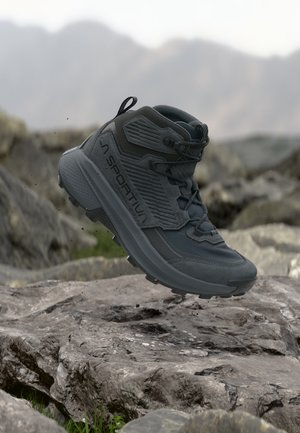 Botte de randonnée gris foncé avec une semelle épaisse flottant au-dessus d’un terrain rocheux, montrant un design robuste et le logo de la marque outdoor La Sportiva.