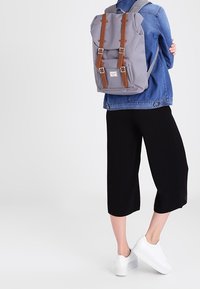 Harmaa reppu, jossa on ruskeat nahkahihnat ja yksityiskohdat. Varustettu yläkannella, säädettävillä hihnoilla ja rennolla muotoilulla. Kannan takana.