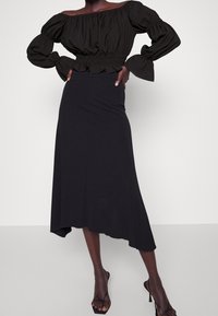 Femme portant un haut noir décolleté avec des manches bouffantes et une jupe midi asymétrique noire à taille haute avec des sandales à talons noires.