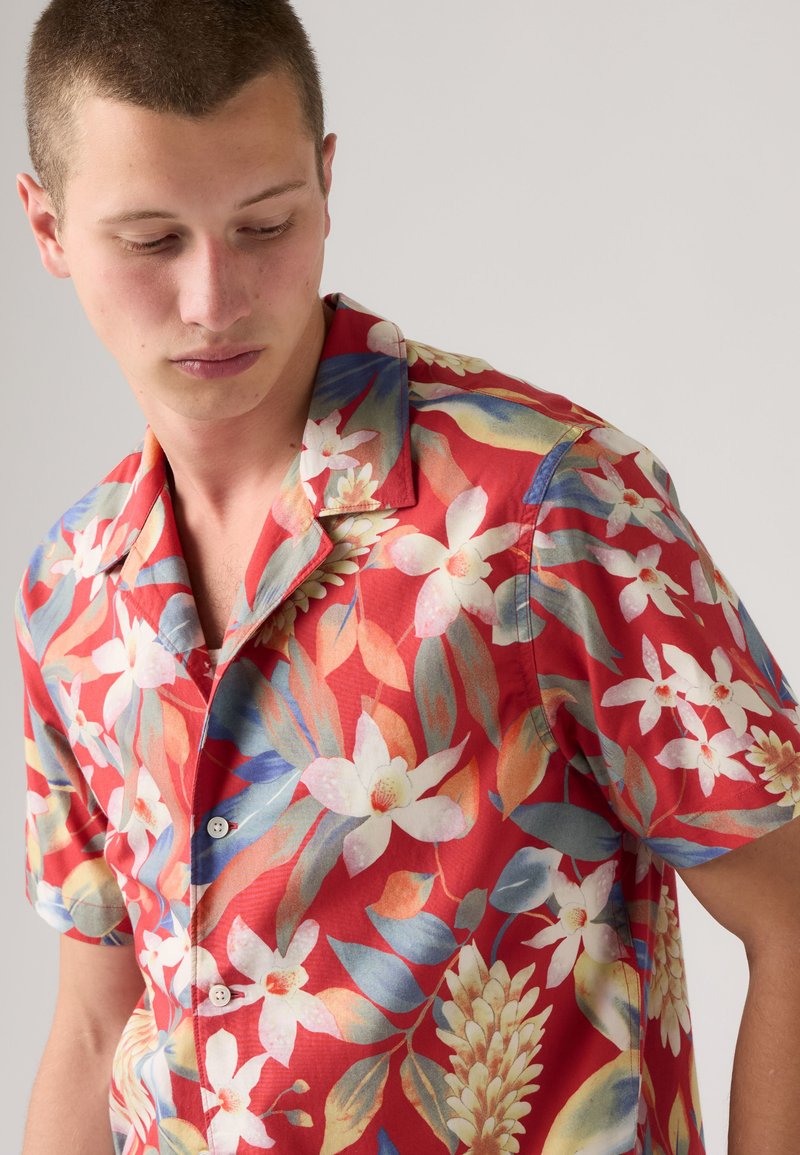 Joven mirando hacia abajo, lleva una camisa roja de manga corta con patrones florales y de hojas en blanco y azul.