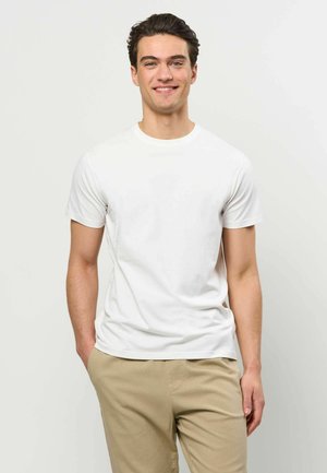 Tânăr zâmbitor, purtând un tricou alb simplu cu mânecă scurtă și pantaloni bej, stând în fața unui fundal simplu, deschis la culoare.