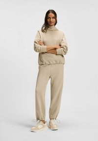 Beige hoodie med framficka och elastiska ärmslut, i kombination med matchande joggers som har elastiska fotledsband, avslutat med beige sneakers.