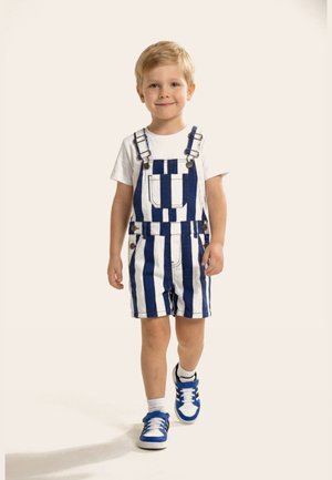 Niño pequeño caminando hacia adelante con una camisa blanca y peto corto a rayas azul y blanco con zapatillas azules y calcetines blancos.