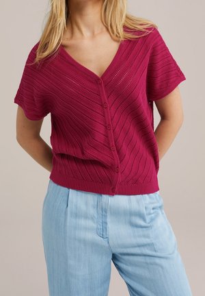 Vrouw draagt een kort mouwtjes, knoopjes top van dieproze gebreide stof met een V-hals, gecombineerd met lichtblauwe broek, staand met handen achter de rug.