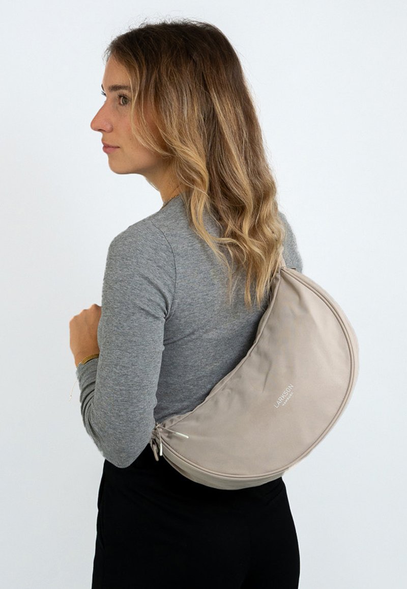 Donna con lunghi capelli ondulati che indossa un top grigio a maniche lunghe e pantaloni neri, mentre porta una borsa beige a forma di mezzaluna sulla schiena.
