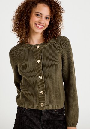 Frau mit lockigem Haar lächelt, trägt eine olivgrüne Strickjacke mit goldenen Knöpfen und schwarze Jeans, steht vor einem schlichten, hellen Hintergrund.