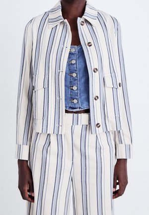 Persoon die een crèmekleurig jack en broek draagt met verticale marineblauwe strepen en daaronder een blauwe denim cropped top met knopen.