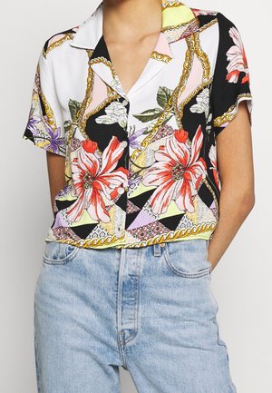 Koszula z krótkim rękawem zapinana na guziki z dużym czerwono-białym kwiatowym wzorem i złotym łańcuchowym motywem, zestawiona z jasnoniebieskimi jeansami z wysokim stanem.