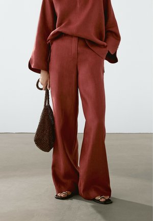 Wide-leg, rust-colored linen pants with a smooth texture. Paired with a black beaded handbag and flat sandals, set against a neutral background.