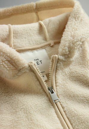 Cremfarbener Reißverschluss-Fleece mit weicher, strukturierten Außenseite, gefütterter Kapuze und silbernem Reißverschlussakzent. Etikett innen sichtbar.