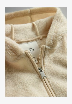 Krémová fleecová mikina na zips s mäkkým, textúrovaným povrchom, podšívkou kapucne a strieborným zipsom. Značka je viditeľná vo vnútri.