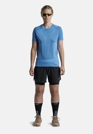Vrouw draagt blauwe sportshirt met korte mouwen, zwarte korte broek, zwarte kniekousen, beige hardloopschoenen en gele sportzonnebril, staand.