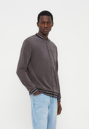 Joven de pie con una mano en el bolsillo de unos vaqueros azul claro, vistiendo una chaqueta gris abotonada con cuello, puños y dobladillo a rayas.