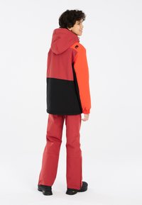 Rote und schwarze Jacke mit Kapuze, elastischen Bündchen; kombiniert mit roten Hosen und schwarzen Stiefeln. Glattes Material, das für Outdoor-Aktivitäten konzipiert ist.