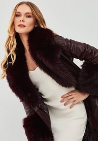 Cappotto in pelle bordeaux con colletto e polsini in pelliccia sintetica, caratterizzato da una rifinitura interna in paisley intricato. La texture è liscia con una finitura lucida.