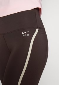 Černé legíny Nike mají těsný střih, s světle béžovým akcentovým pruhem po straně a bílým logem "AIR" v oblasti boků.