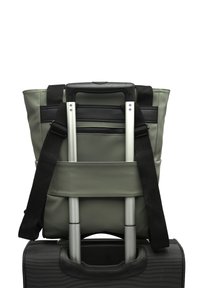 Borsa a tracolla grigia con dettagli neri, appoggiata su una valigia nera con ruote. Dotata di spallacci regolabili e di una tasca posteriora con cerniera.