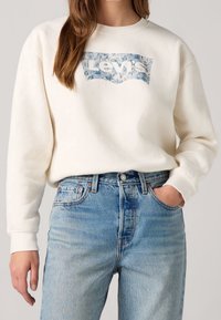 Sudadera color crema con un logotipo de Levi's en azul con flores. Combinada con pantalones vaqueros de corte alto y de un tono azul claro, de ajuste relajado y diseño clásico.