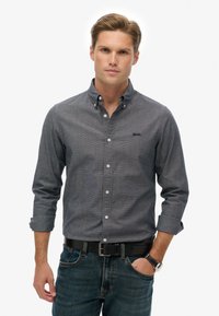 Langærmet, knap-ned skjorte i mørkegrå med et subtilt mønster. Har en krave, knaplukning og brystlogo. Bæres med blå jeans.