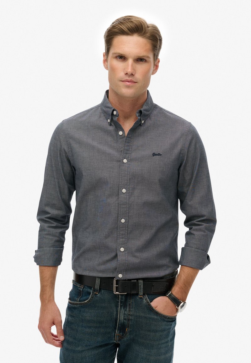 Langærmet, knap-ned skjorte i mørkegrå med et subtilt mønster. Har en krave, knaplukning og brystlogo. Bæres med blå jeans.