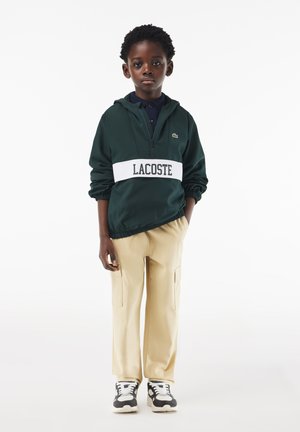 Giacca verde con cappuccio e cintura bianca che presenta il logo "LACOSTE", abbinata a pantaloni cargo beige e sneakers bianco e nere.