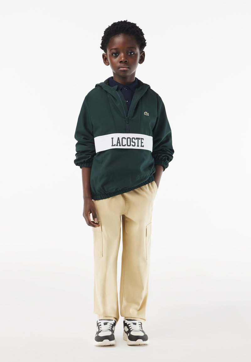 Green hooded jacket with white waistband featuring "LACOSTE" logo, paired with beige cargo pants and black-and-white sneakers.