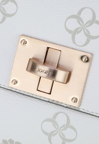 Gros plan sur une boucle de sac à main rectangulaire de couleur dorée avec l'inscription "JOOP!", fixée à un cuir blanc orné de motifs floraux gris clair.