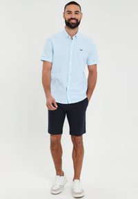 Camicia button-up a maniche corte azzurro chiaro con colletto e piccolo dettaglio ricamato, abbinata a pantaloni corti blu marino e sneakers bianche.