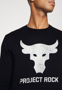 T-shirt noir à manches longues avec un grand logo de taureau blanc craqué et le texte "PROJECT ROCK" en lettres blanches grasses en dessous. Texture de tissu doux.