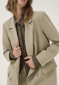 Beige tekstureret blazer med spidse lapels, to store knapper og forlommer, lagdelt over en mørk, blomsterprintet skjorte.