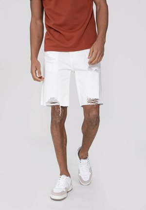 Shorts en denim blanc avec des bords effilochés et des détails déchirés. Associés à un t-shirt marron et des baskets blanches avec des accents gris.