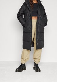 Černý péřový kabát s kapucí, s prošíváním ve tvaru čtverců a zapínáním na zip. Kombinován s khaki kalhotami a černými koženými kotníkovými botami na šněrování.