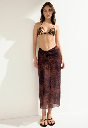 Vrouw staat tegen een witte muur, draagt een bikinitop met tijgerprint, een donkerbruine sarong met luipaardprint en zwarte sandalen met gouden kettingdetail.